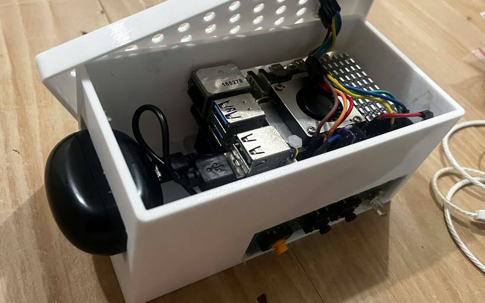 The EarPrompt enclosure with its lid open: a Raspberry Pi 5 with an active cooler, colour-coded wiring running to the lid, and a black Bluetooth earbud case docked on the side.