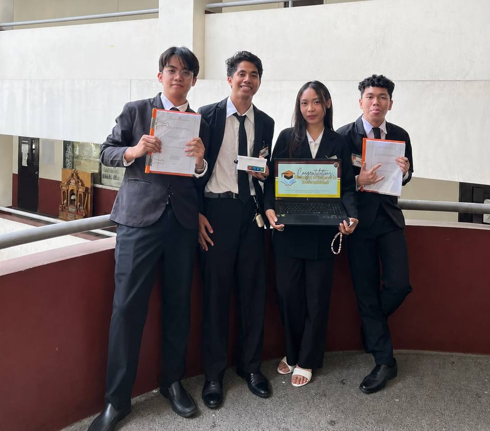 The four EarPrompt researchers in business attire after their thesis defense, holding bound manuscripts, the EarPrompt device and a laptop with a congratulations slide.